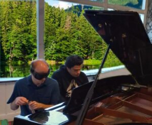 Sky and Alex, two young blind men, sharing a piano bench at a grand piano with its lid open. Sky is to the left. He is slightly bald, wearing learning shades and holding a travel mug. To his right, Alex is looking down while playing during a concert. Both have light skin and dark brown hair, and they are both wearing dark blue shirts. In the background, the sun bathes the trees of a nearby mountain on a sunny day.