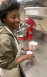 Kadija, who has dark curly hair and beautiful dark skin smiles at the camera while holding a package of butter. She is wearing a military green shirt with a white apron. Kadija is standing in front of a stainless steel kitchen counter, where a white and red bowl and a red stand mixer are visible in the background.