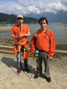Alex and Aedan standing, posing for the camera. Alex is wearing his staff shirt with a safety jacket tied around his waist. Aedan is also wearing his staff shirt overtop of which he is wearing an unzipped orange rain jacket. Both Alex and Aedan are holding their canes.